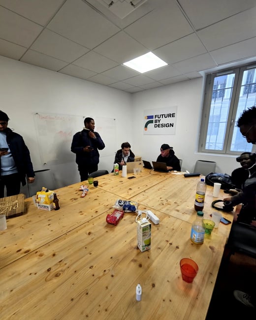 A group of people are gathered around a large wooden table in an indoor setting, some using laptops while snacks and drinks are scattered across the surface.