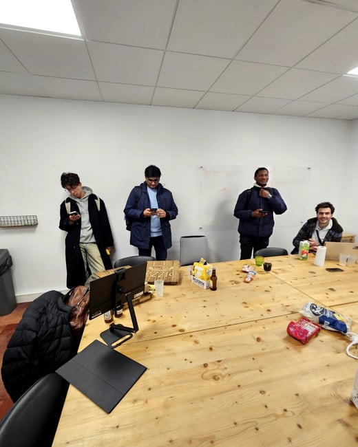 A group of four young men are in a room with a large wooden table, some looking at their phones while others pose for the camera. The table is cluttered with pizza boxes, drinks, and snacks, suggesting a social gathering or a break.