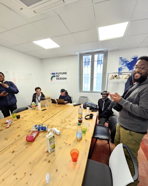 A group of five men are gathered in a bright office space around a large wooden table with laptops, snacks, and drinks.