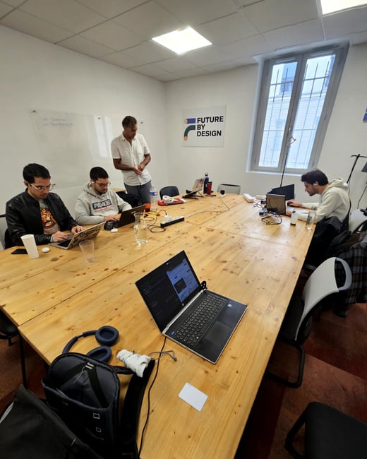 Four men are working on their laptops around a large wooden table in an office environment, with a 'FUTURE BY DESIGN' logo visible on the wall.