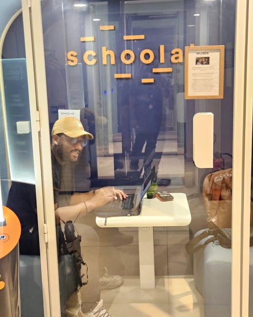 A man wearing a yellow cap and glasses sits inside a glass-enclosed 'schoolab' workspace booth, focused on his laptop.