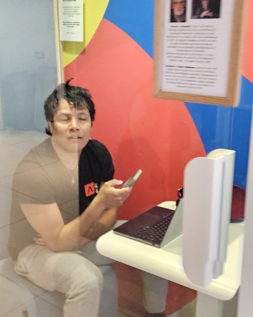 A person sits in a colorful workspace booth, looking down at a smartphone with their eyes closed or blinking, next to an open laptop.