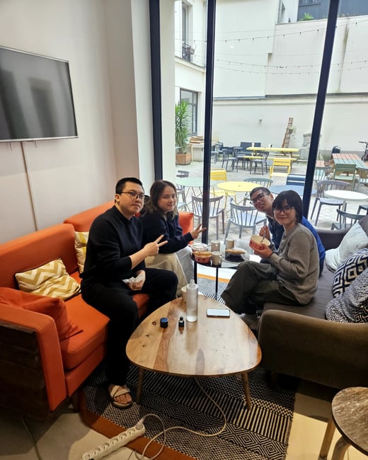 A group of four young adults is sitting in a modern lounge area, posing for a photo while sharing a meal around a coffee table.