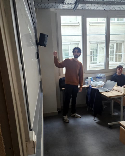 A young man in a brown sweater stands next to a whiteboard, pointing towards it while another person sits at a desk with a laptop in the background.