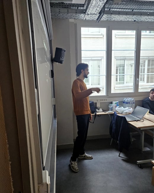 A man in an orange sweater stands in a room, seemingly giving a presentation or explanation next to a whiteboard and a desk with a laptop.