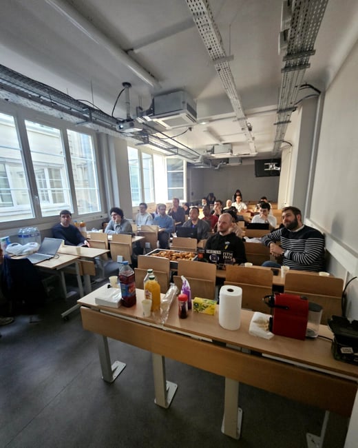 A group of people are seated in a classroom or lecture hall, facing forward towards the camera, with a table of snacks and coffee in the foreground.