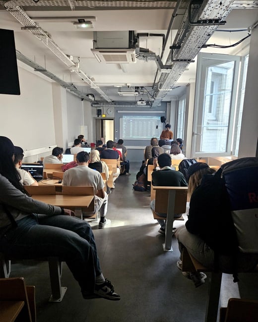 A group of students sitting in a classroom or lecture hall, attending a presentation shown on a screen at the front of the room.