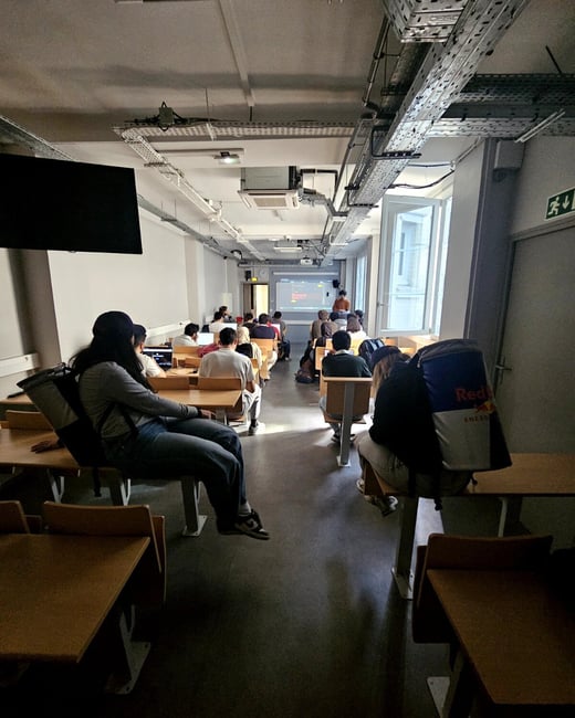 A group of students are seated in a classroom or lecture hall, focused on a presentation being displayed on a screen at the front of the room.