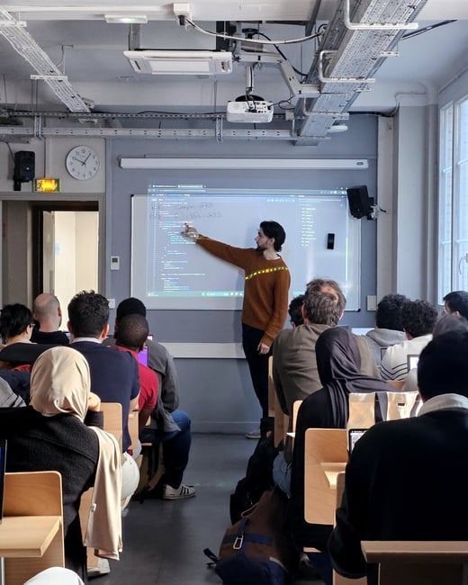 A man stands at the front of a classroom, pointing at a projected screen displaying code while an audience of students watches.