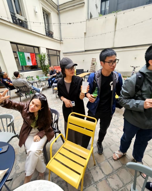 A group of people are socializing in an outdoor courtyard area with tables, chairs, and a Mexican flag hanging in the background.