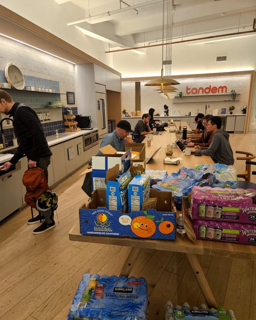 An indoor office or co-working space featuring a kitchen area and a long communal table where several people are working on laptops. In the foreground, there are various boxes and packs of snacks and beverages, including Sunkist mandarins and Kirkland water.