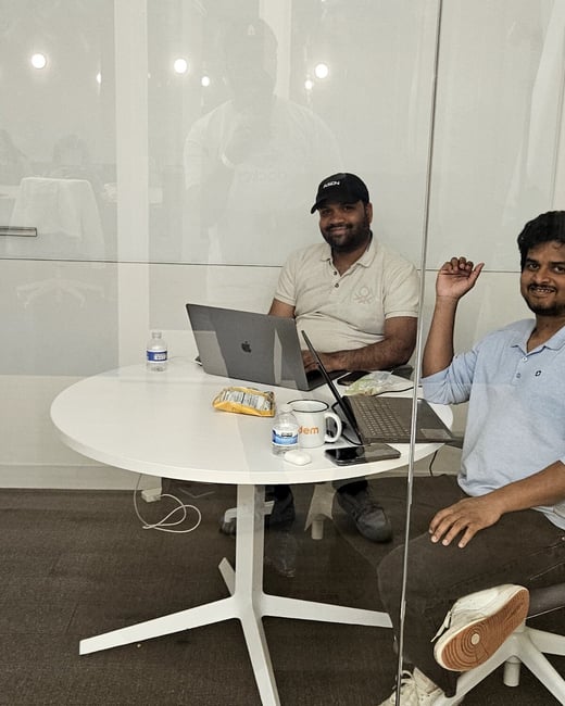 Two men are sitting at a round white table in an office setting, both looking towards the camera while working on their laptops.