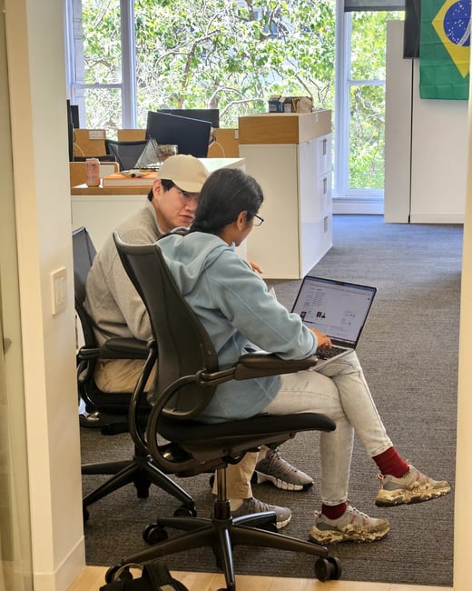 Two people are sitting in an office environment, focused on a laptop screen together in a collaborative manner.