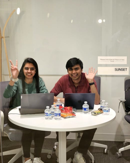 Two young adults, a woman and a man, are sitting at a round white table in an office or study space, smiling and waving at the camera with laptops and snacks in front of them.
