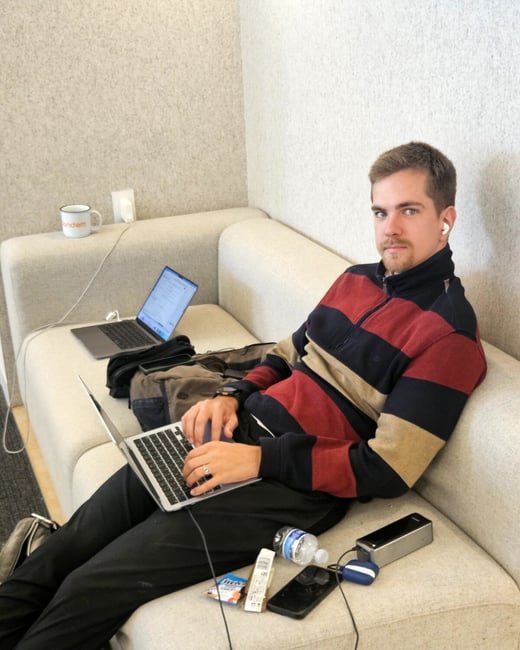 A man is sitting on a light-colored couch, working on a laptop while looking directly at the camera.