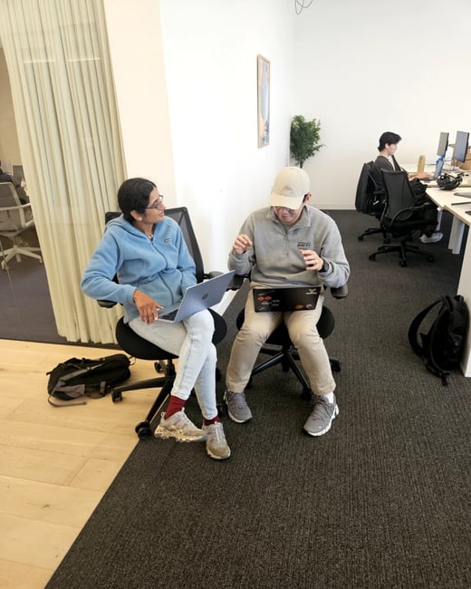 A woman and a man are sitting in office chairs with laptops, engaged in a discussion within a modern office setting. Another person is visible working at a desk in the background.