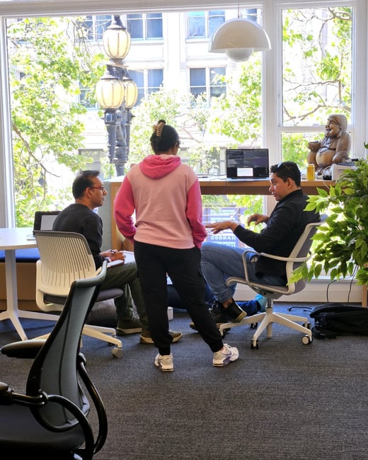 A group of three people are engaged in a discussion within a bright, modern office space featuring a large window with a city view.