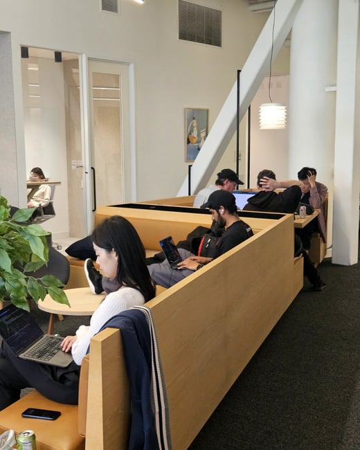 A modern, open-plan co-working space where several individuals are focused on their laptops or engaged in quiet work.