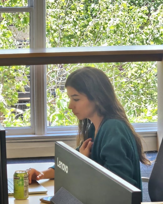 A woman with long brown hair is sitting at a desk working on a computer, positioned in front of a large window with a view of lush green trees.