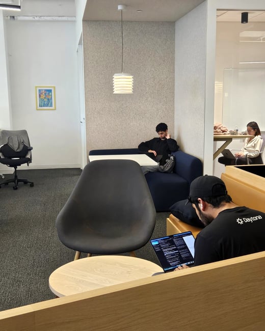 The image shows a modern, minimalist office or co-working space where several people are focused on their laptops.