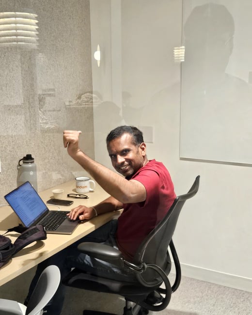 A man in a red t-shirt sits at a desk in an office, smiling and raising his fist in a celebratory gesture while looking at the camera.