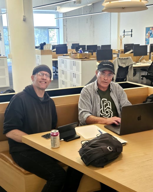 Two men are sitting at a wooden table in a modern open-plan office; one is smiling at the camera while the other works on a laptop.