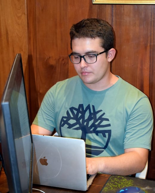 A young man with glasses and a green t-shirt is working on a laptop at a wooden desk, with an external monitor visible to the left.