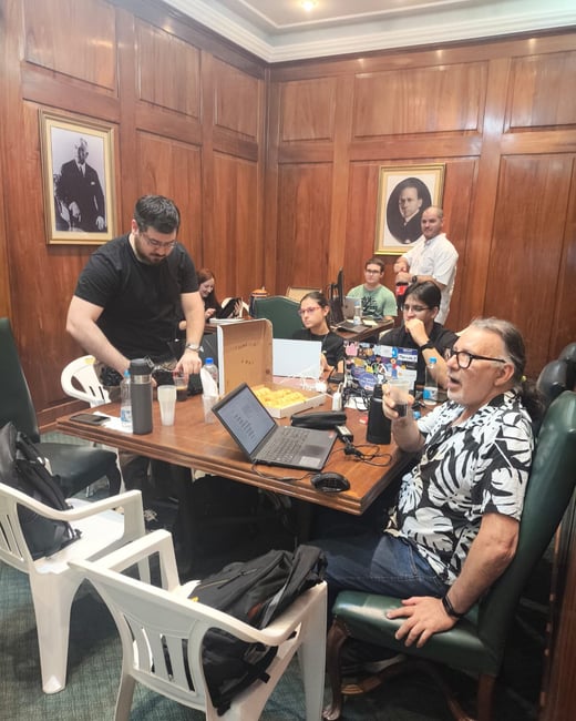 A group of people are gathered around a large wooden table in a wood-paneled room, appearing to be in a meeting or a collaborative work session with laptops and snacks.