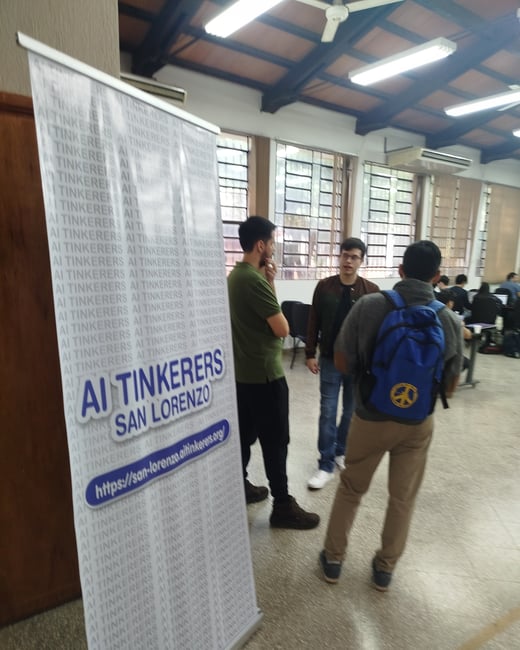 A photograph of an indoor event featuring a prominent roll-up banner for 'AI TINKERERS SAN LORENZO' with several people standing and talking in the background.