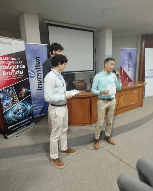 Three men are on a stage during an event, with one man speaking into a microphone while the others stand nearby next to banners about Artificial Intelligence.