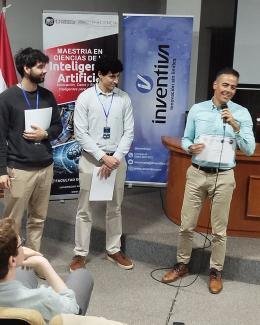 A man speaks into a microphone while holding a certificate on a stage, standing next to two other men during an academic or professional event.