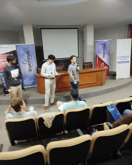 A presentation event taking place in an auditorium, featuring three speakers on a stage and an audience seated in the foreground.