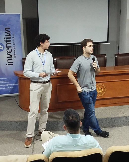 Two men are on a stage in an auditorium during a presentation; one is speaking into a microphone while the other stands by holding papers.