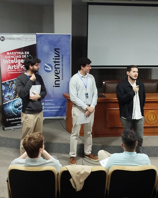 Three men are standing on a stage during a presentation, with one man speaking into a microphone. Behind them are banners for a Master's in Artificial Intelligence and a company called Inventiva.