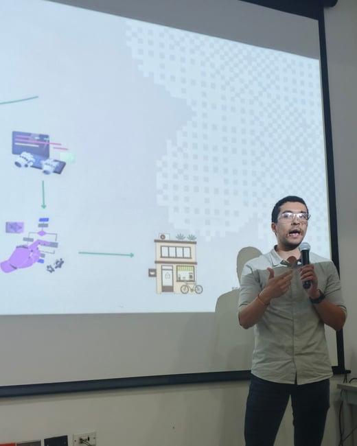 A man is giving a presentation, holding a microphone and gesturing in front of a large projection screen displaying a diagram.