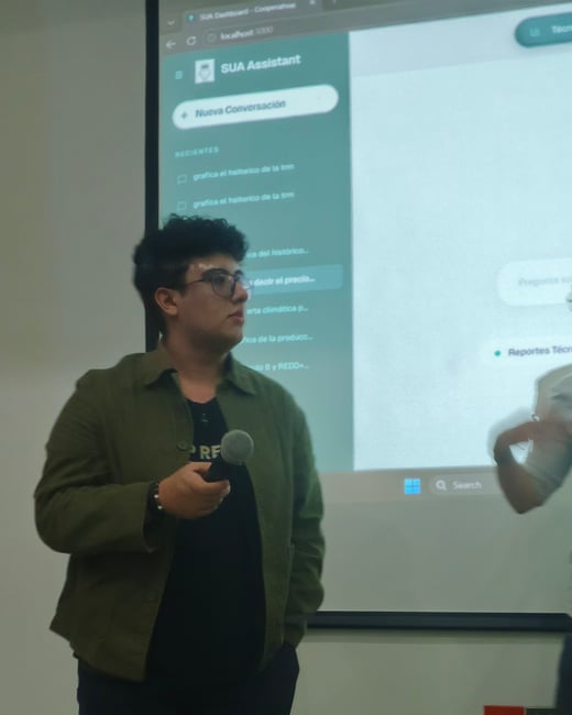 A person wearing glasses and a green jacket holds a microphone while giving a presentation in front of a projection screen displaying a web application.