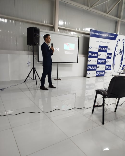 A man in a blue suit stands in front of a projector screen and a banner, speaking into a microphone during a presentation in a room with a reflective tiled floor.
