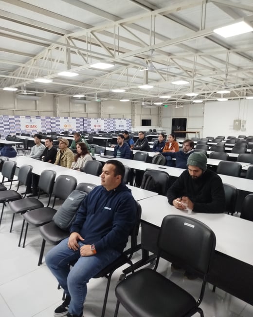 An indoor photograph of an audience seated at rows of white desks in a large lecture hall or warehouse-like classroom, listening to a presentation.