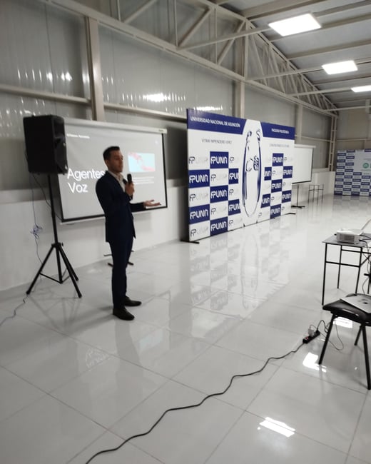 A man in a dark suit is giving a presentation, holding a microphone in front of a projector screen displaying 'Agentes Voz' inside a large hall with a white tiled floor and a university backdrop banner.