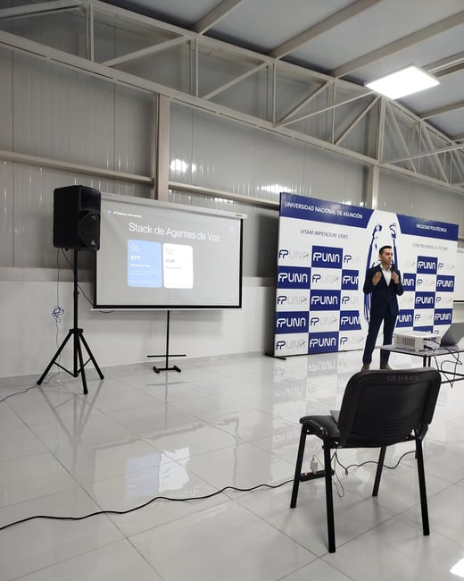 A man in a suit is giving a presentation in a large hall, standing next to a projector screen displaying a slide about voice agent stacks and in front of FPUNA university banners.