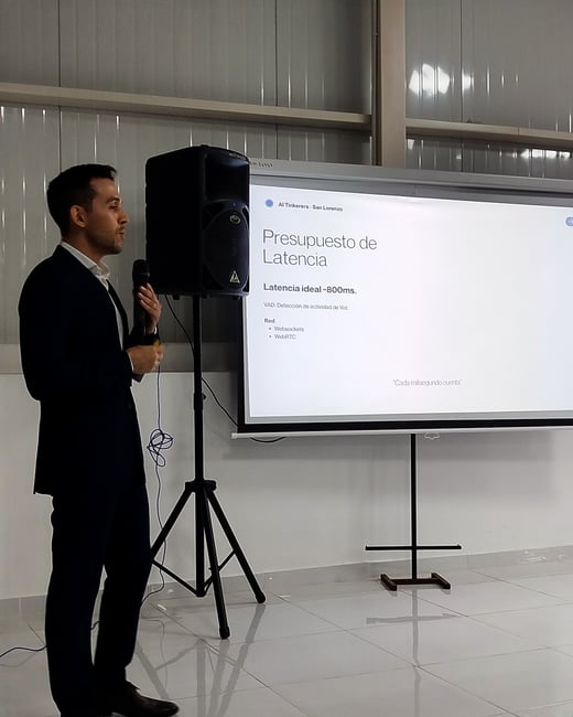 A man in a dark suit stands indoors holding a microphone, delivering a presentation next to a projector screen displaying a slide about latency budget in Spanish.