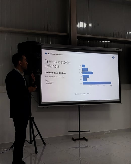 A man in a dark suit stands next to a projection screen presenting a slide about latency budgets in Spanish.