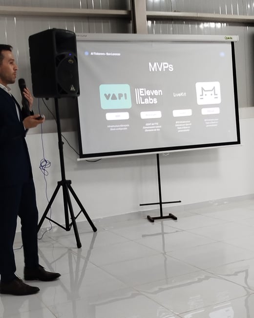 A man in a dark suit is giving a presentation next to a projector screen displaying various AI MVPs like Vapi, ElevenLabs, LiveKit, and Ollama.