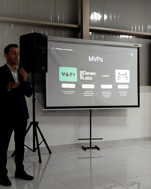 A man in a dark suit is standing next to a projector screen, delivering a presentation about MVPs in AI technology, featuring logos like VAPI, ElevenLabs, and LiveKit.