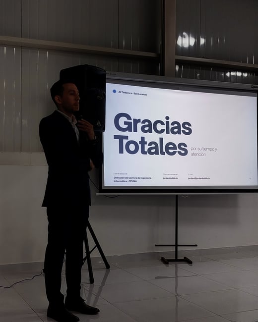 A man in a dark suit stands next to a projection screen displaying a presentation slide that reads 'Gracias Totales'.