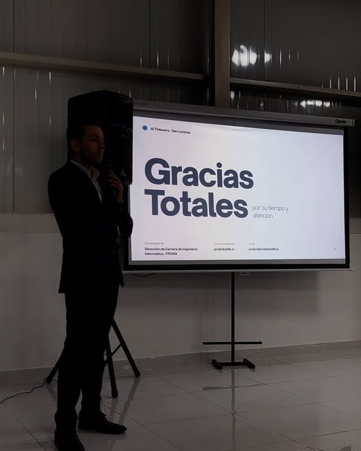 A man in a dark suit stands next to a projector screen displaying a presentation slide that says 'Gracias Totales' in Spanish.