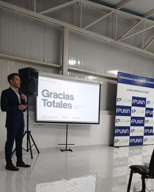 A man in a blue suit stands next to a projector screen displaying 'Gracias Totales' during a presentation, with an FPUNA banner in the background.