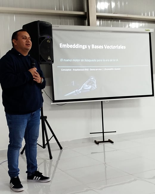 A man stands indoors next to a projection screen, delivering a presentation about embeddings and vector databases in Spanish.