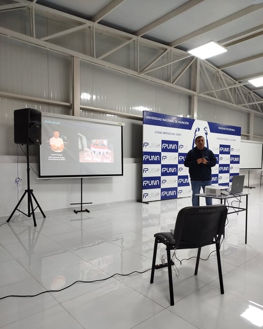 A man stands presenting in a large hall next to a projector screen and a promotional banner for the Universidad Nacional de Asunción.