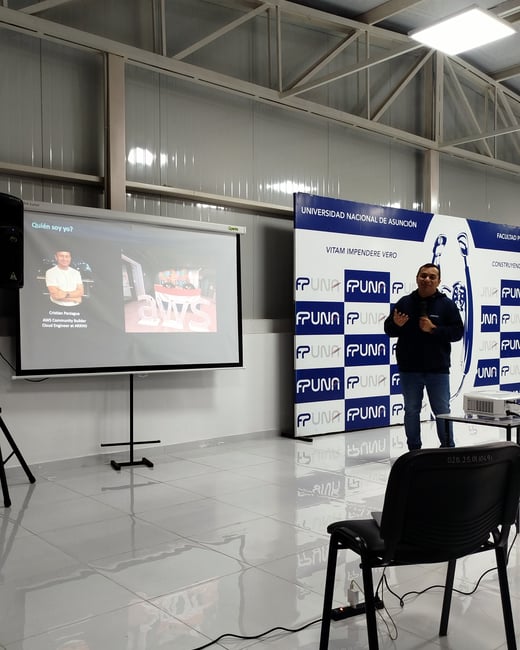 A man stands presenting in front of a projector screen and a branded backdrop in a large room with a tiled floor.